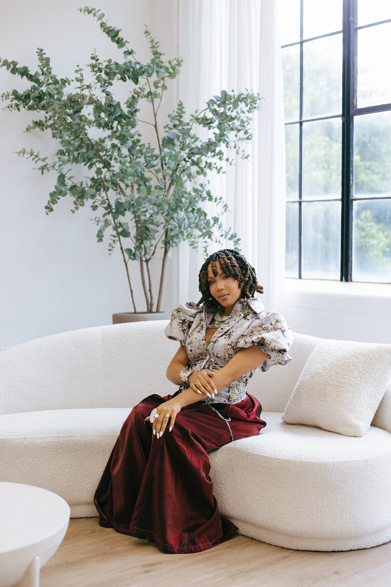The woman behind House of Dvine, seated in a bright room in a deep oxblood satin skirt